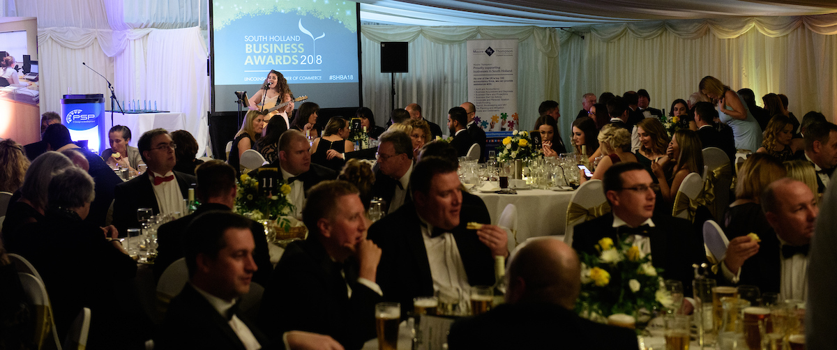 Group of people in eveningwear standing and talking at South Holland Business Awards 2018 with singer on stage