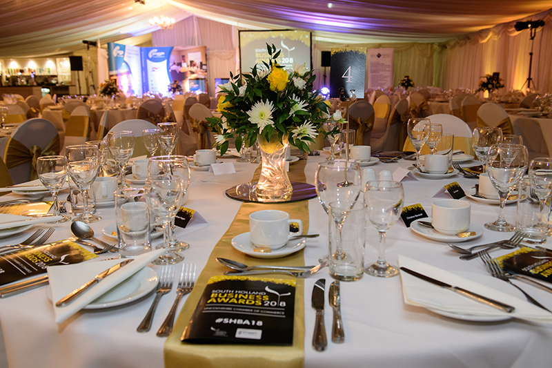 Close up of a table with white tablecloth with cutlery, glassware, floral centre piece and black booklet in a room at an awards show