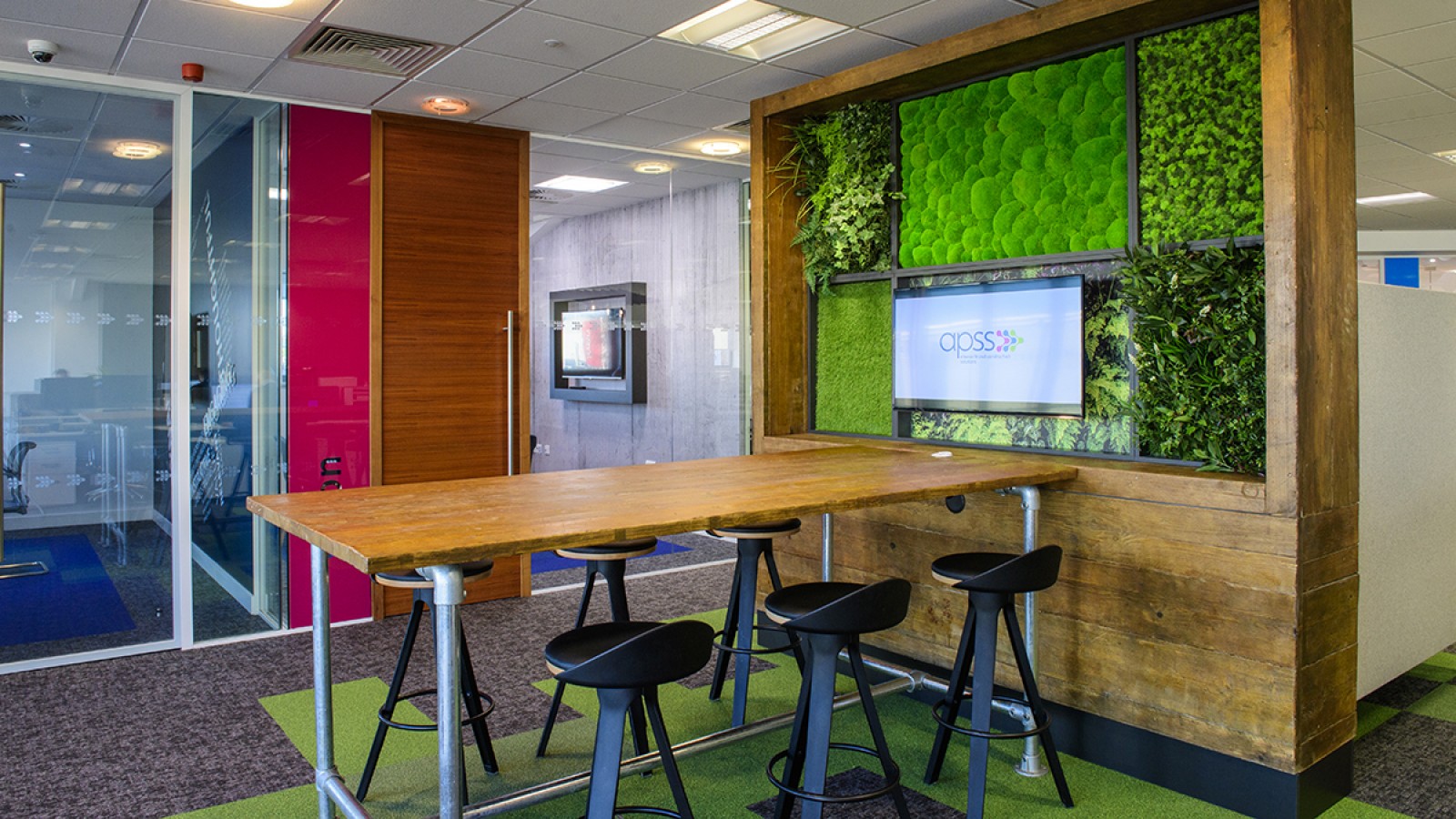 The interior of a newly refurbished office, with high table, chairs, feature wall with greenery, and screen showing APSS logo