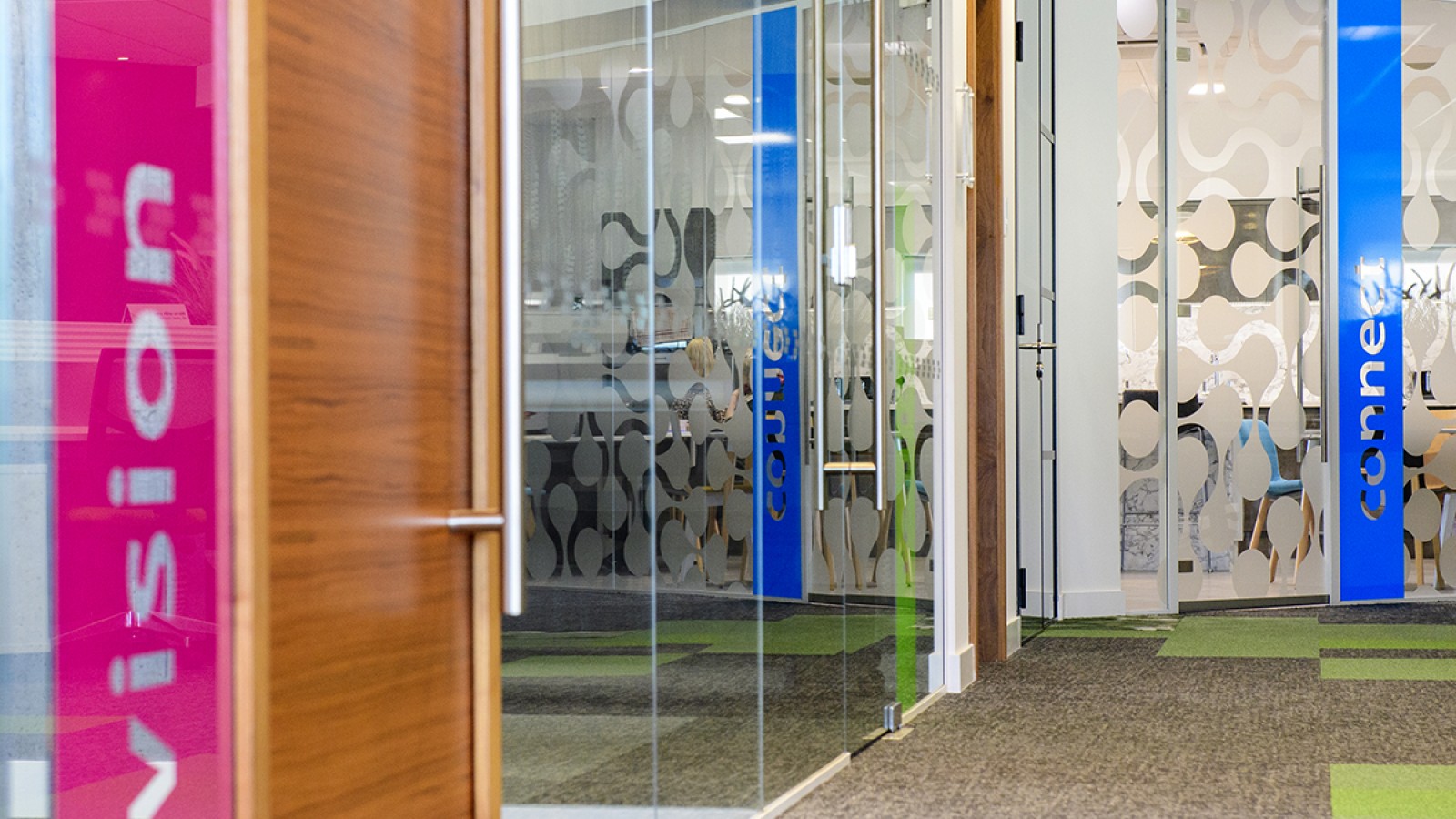 The interior of a newly refurbished office, with glass walls and doors, grey and green carpet and green and blue signage
