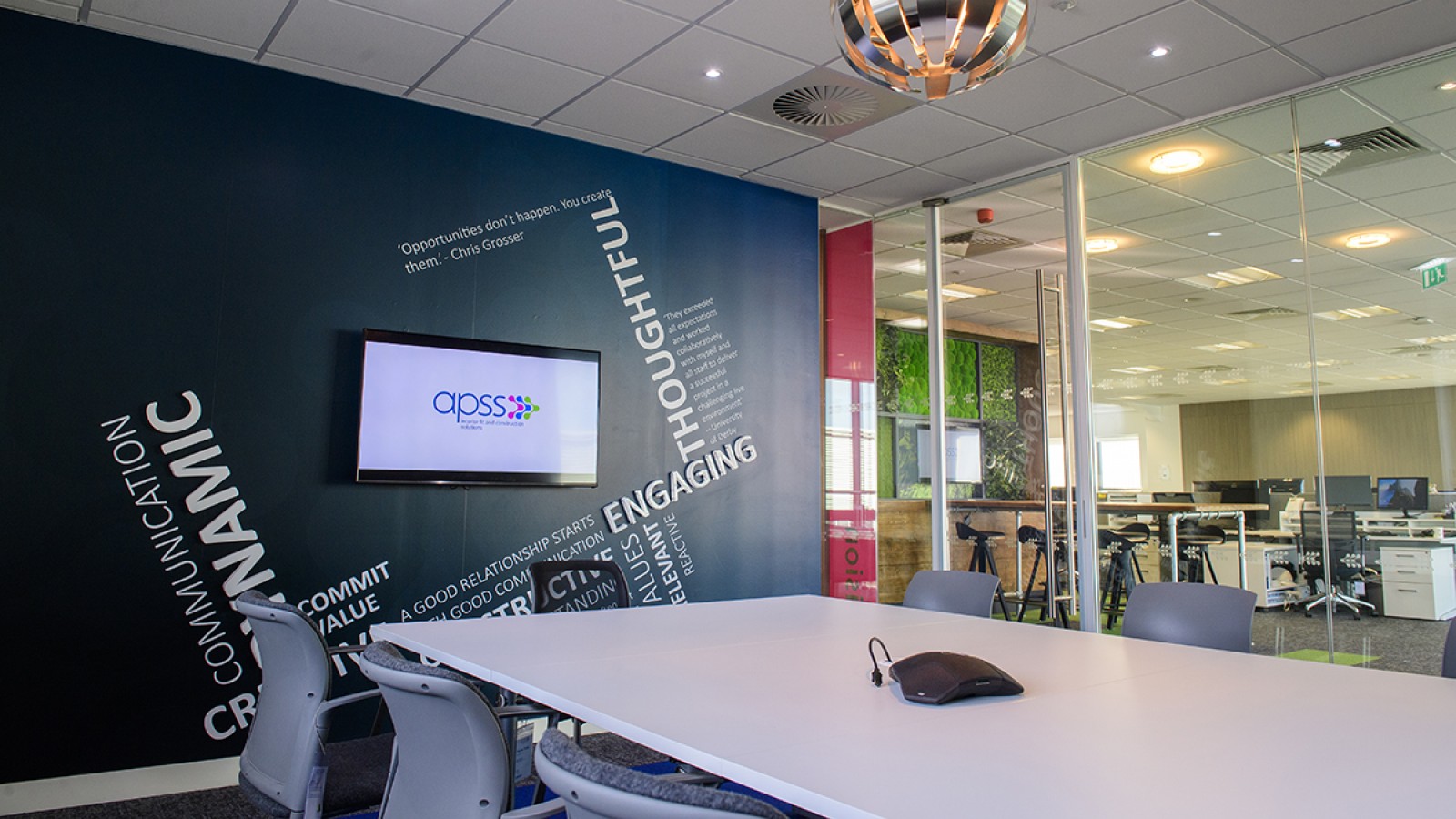 The interior of a newly refurbished office, with a table, chairs, feature wall with text, and screen showing APSS logo