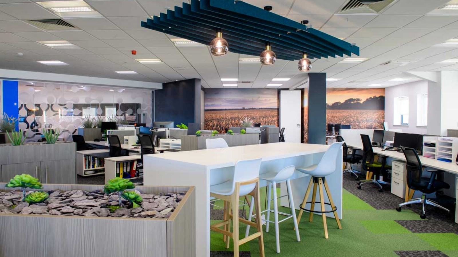The interior of a newly refurbished office, with desks, high table, chairs, feature wall with mural, plants and green carpet