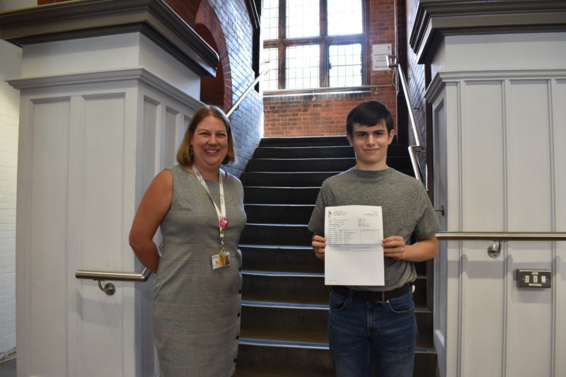 A blonde smiling woman and a young man holding a paper with his GCSE results in front of a staircase