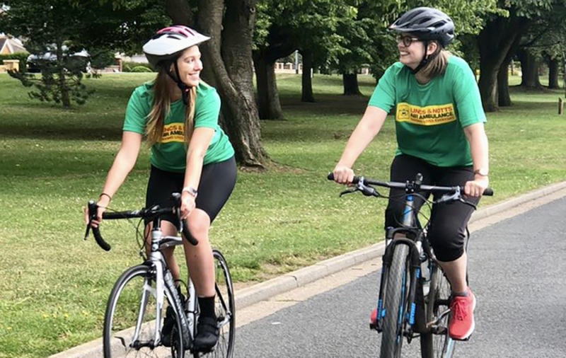 Two young women riding bikes with safety helmets and wearing green T-shirts with theyellow Lincs and Notts Air Ambulance logo