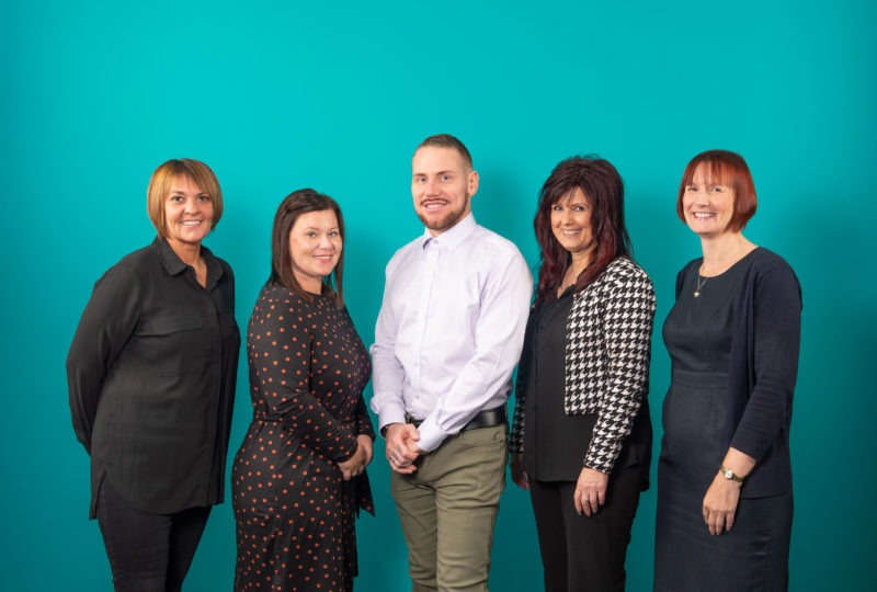 Group of women and men posing in smart casual wear in front of bright teal coloured wall