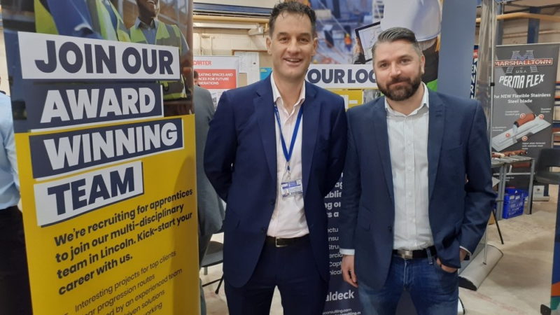 Two white males at exhibition with banner with text "join our award winning team"