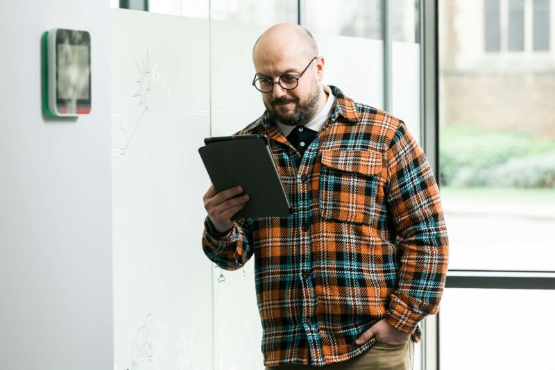Man holding tablet device in his hand, he is wearing orange and grey plaid and spectacles. 