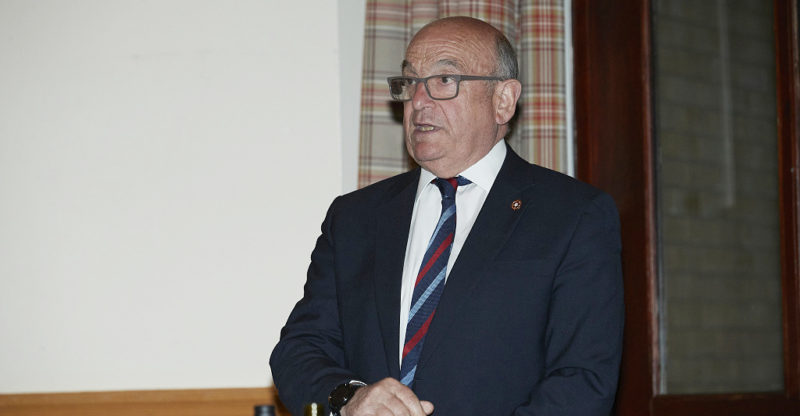 Stuart Peach, a white male wearing dark suit and striped red, blue and white tie with glasses, standing and speaking