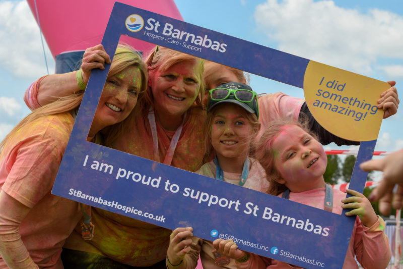 Two women and three children holding a large frame in which they are posing. The blue frame has text "I did something Amazing" and "I am proud to support St Barnabas" and the people are covered in pink and green powder paint.