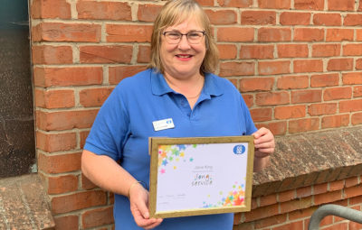 A woman standing in front of a brick wall, wearing a blue Lincolnshire Co-op top, holding a Long Service certificate. 
